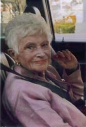 Older Muriel sitting in passenger seat, dressed in pink.