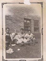 Old faded photo of four young men sitting on lawn outside house.