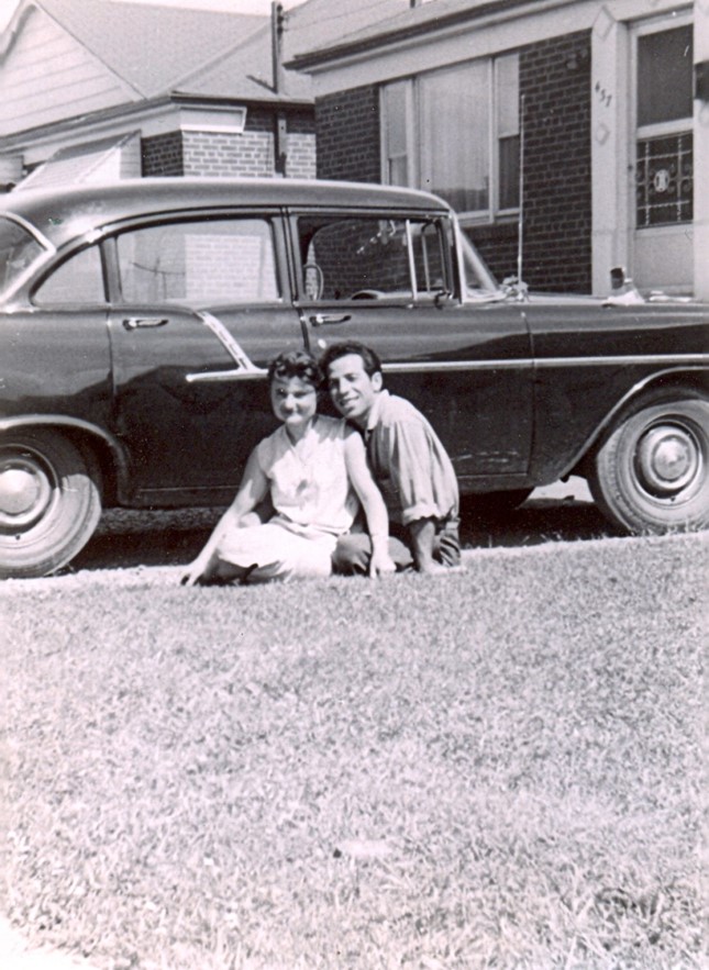 A black and white photo of an Italian man and woman sitting on a driveway with a car behind them.