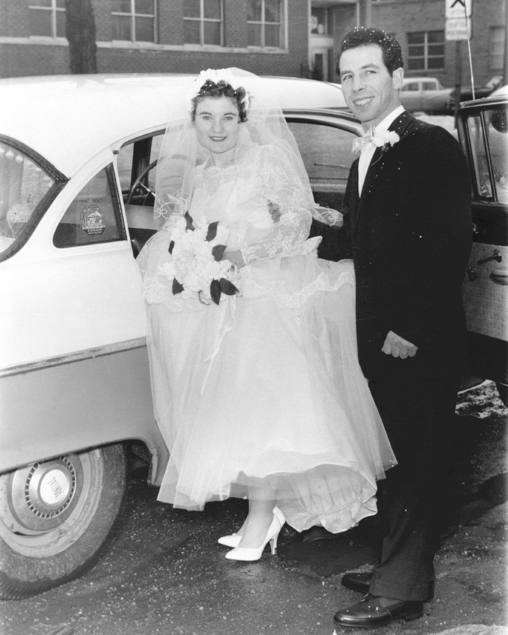 A black and white photo of an Italian man and woman on their wedding day getting into a car.