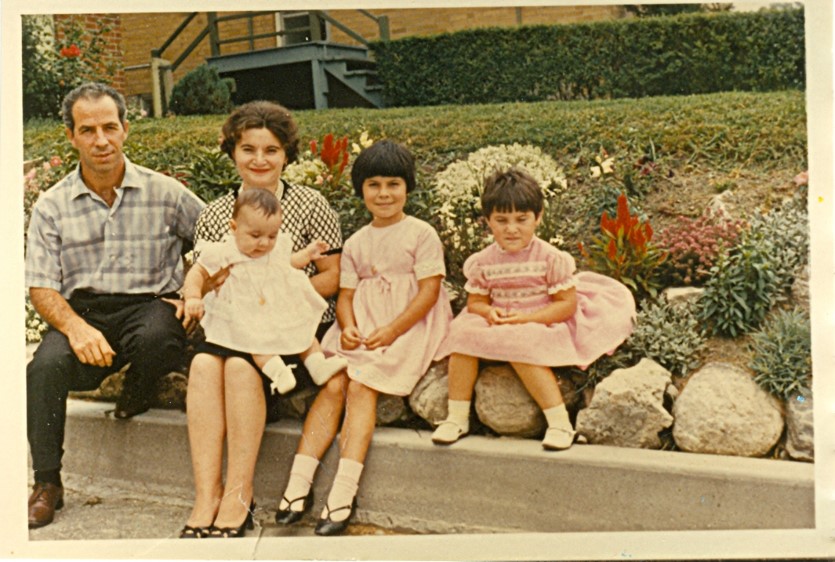 An old photo of an Italian family, including a man, woman, and three children, all dressed nicely and sitting outside.