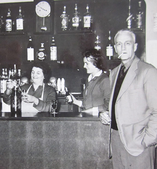 A man stands in front of a bar smoking a cigarette, while two women are behind the bar.