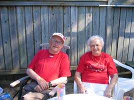 Recent photograph of Noel and Noreen, older, wearing red T-shirts, sitting on the patio.
