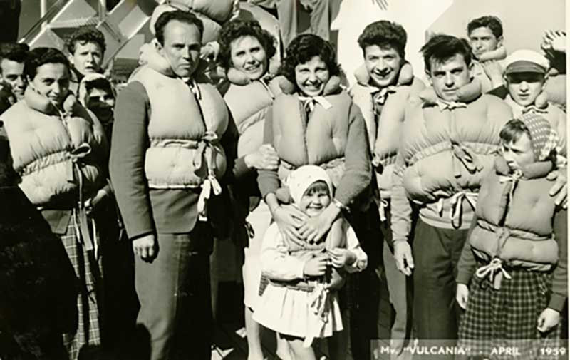 A group of adults and children are standing on the deck of a ship and all are wearing life jackets.