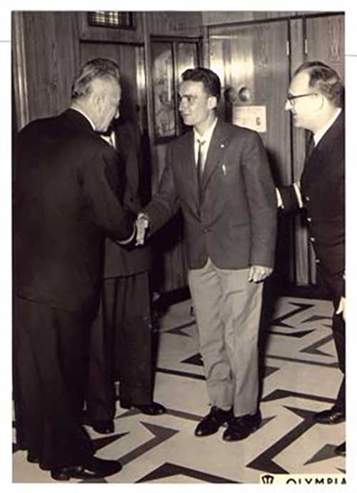 Young man shakes hands with older men while two others look on.