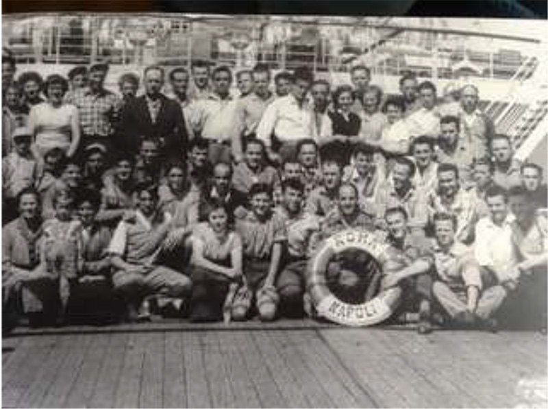 Black and white photo group of people sitting and standing in front of ship.
