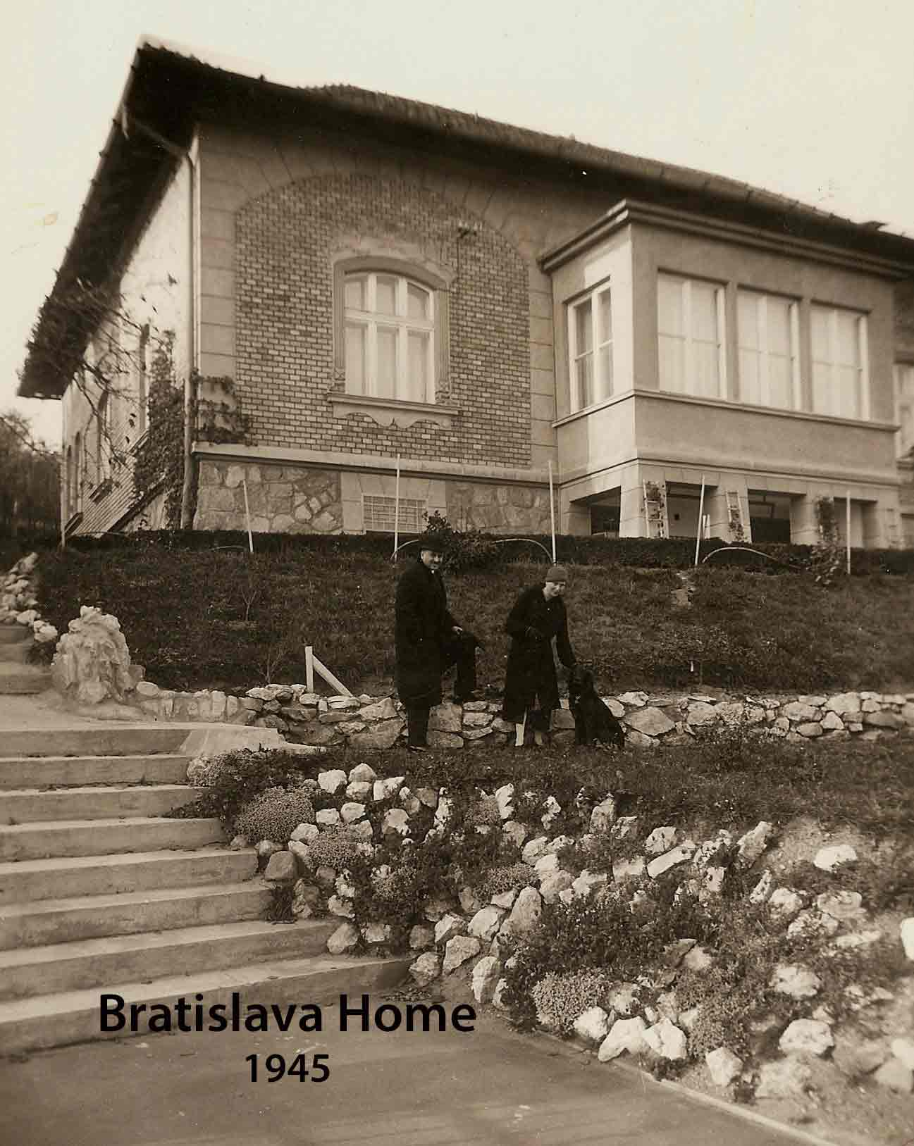 A sepia toned photo of two people and a dog in front of a house with a brick ledge.