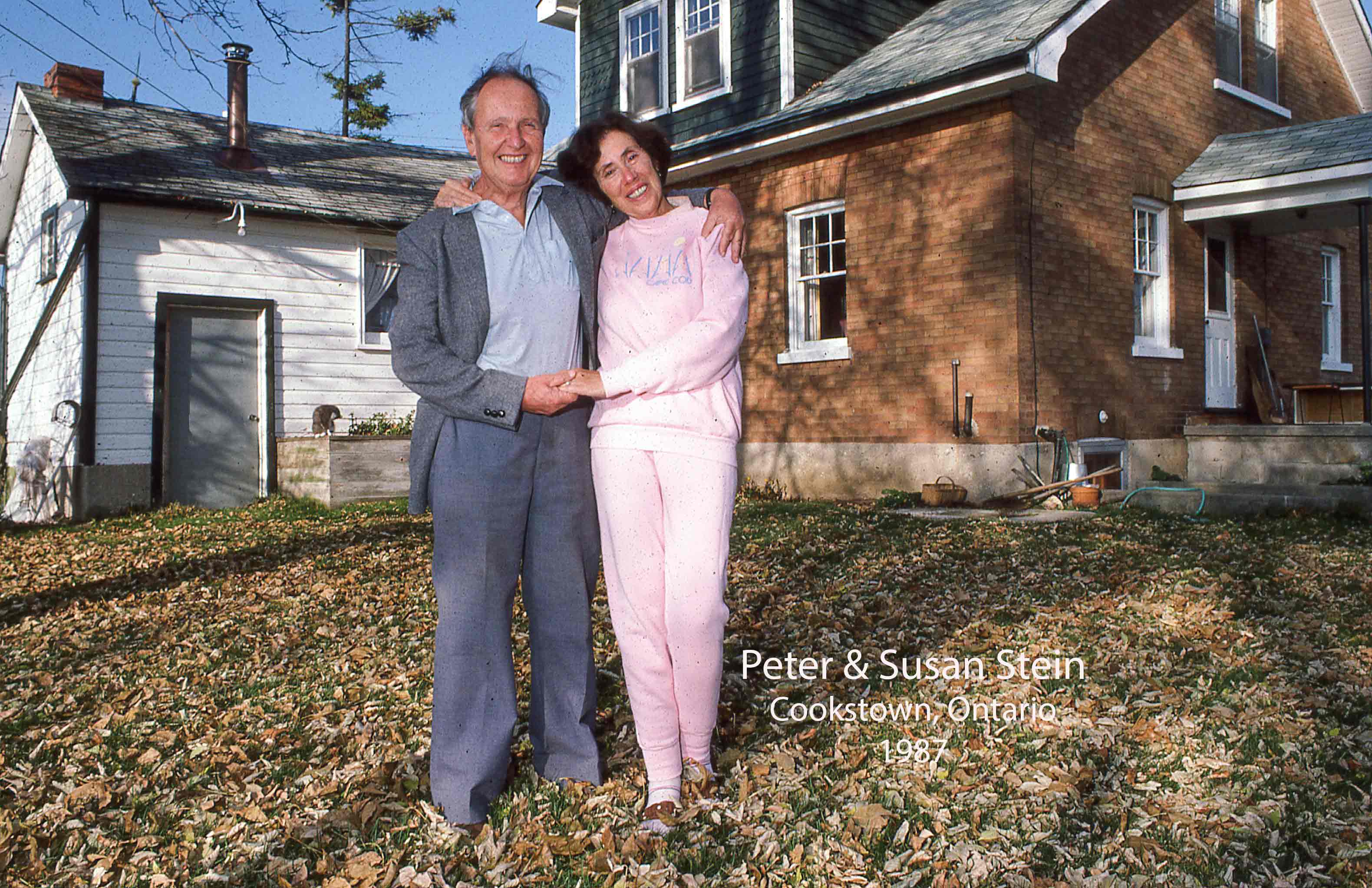 An older Slovakian man and woman standing outside a house in fall leaves, smiling at the camera.