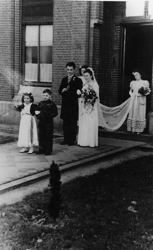 Old photo of married couple coming out of the building. Two kids are walking in front of them and one lady is holding the bridal dress.