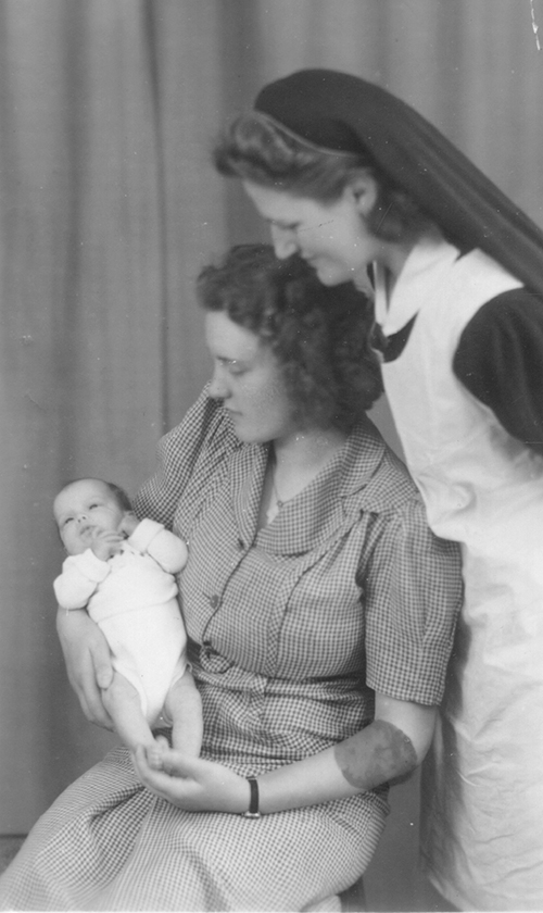 A beautiful lady sitting on a stool holding a new born baby with another woman standing behind her.
