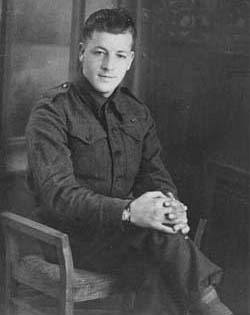 Young man in military dress, seated in chair with hands clasped around one knee.