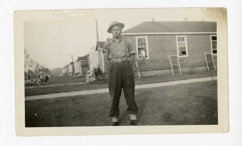 A young man in a military uniform stands on the grass in front of a house.