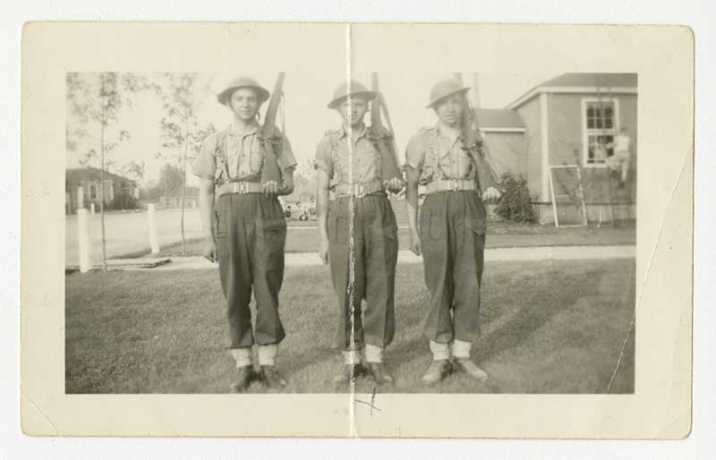 Three young men in military uniforms; all are holding a rifle over their left shoulder.