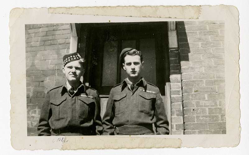 Two young men in military uniform stand in front of a cement building.