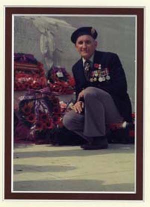 Recent photograph of older Langille, in military dress, kneeling near a wreath of flowers placed near a cenotaph.