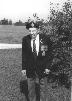 Older Norman, wearing the Legion uniform, sitting on the grass in front of a bush. 