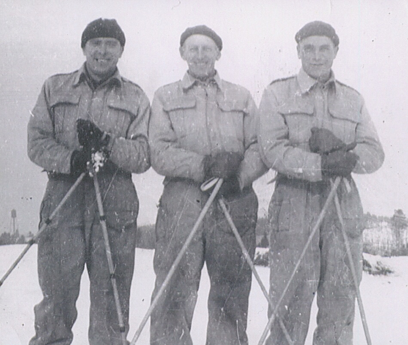 Three men wearing winter clothing stand side by side, each man is holding ski poles.