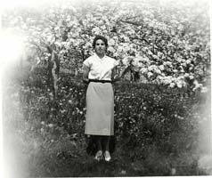 Young woman in skirt and blouse standing in front of trees.