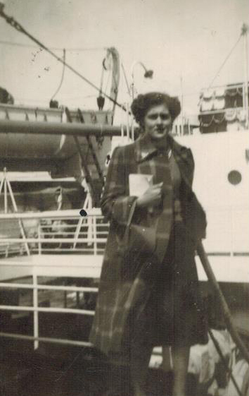 A woman is standing on the deck of the ship wearing a long checkered coat and holding a book in one hand.