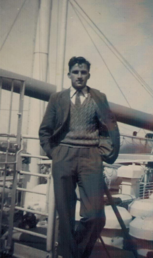 A young man wearing a three-piece suit leans against the railing of a ship and has his hands in his pockets.