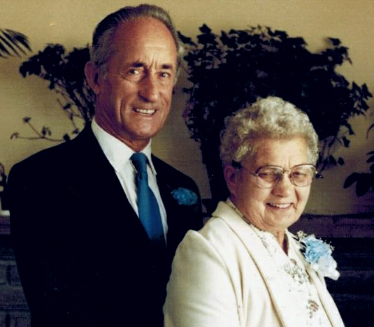 A well-dressed couple smile at the camera, the woman is wearing a blue corsage.