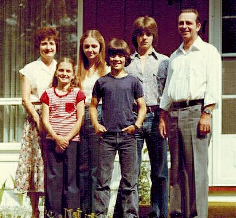 A man and woman stand on the lawn in front of their house, four children stand between them.