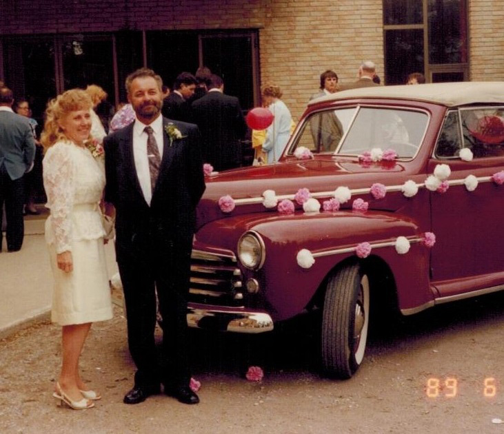 A man and and woman stand in front of an old-fashioned car that has been decorated with paper flowers and streamers.
