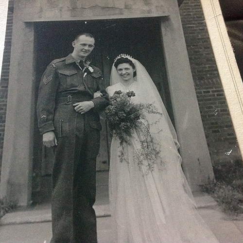 Newly wedded couple is standing in front of building, with the bride holding flowers.