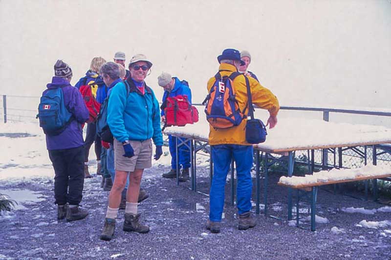 A group of hikers stand in front of an outdoor table, there is snow on it.