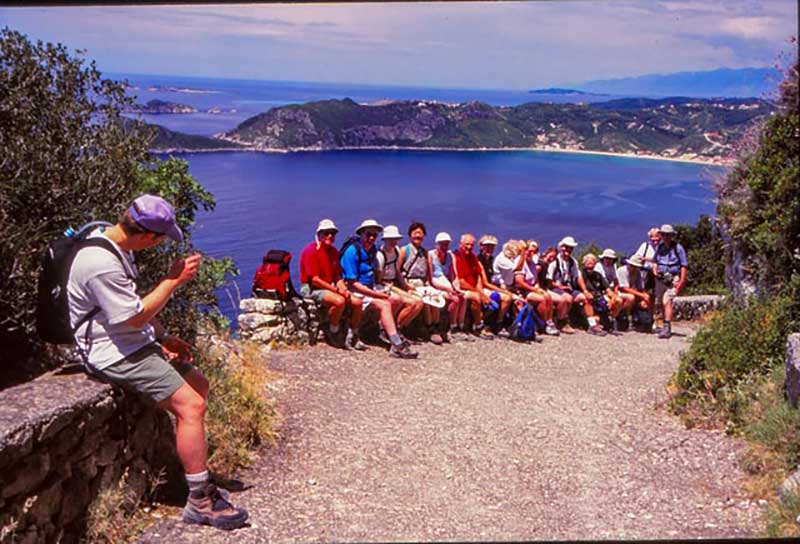 A group of tourists sit in a row on a rock wall, with water and hills behind them.