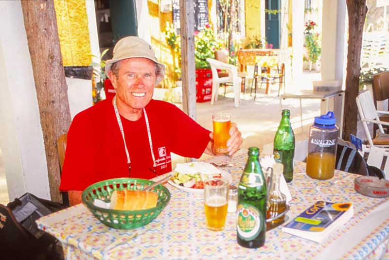 A man in a red t shirt sits at a table laden with food and drink.