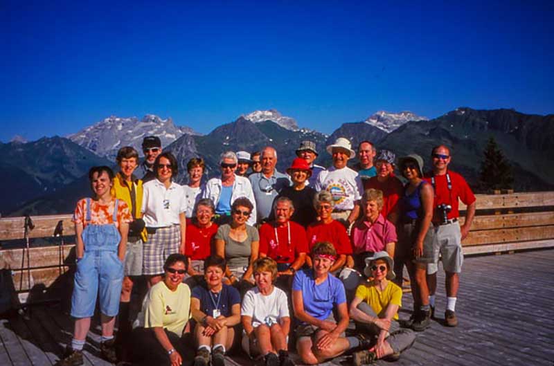 A group of tourists stand and kneel in front of a rock wall, there are beautiful mountains behind them.