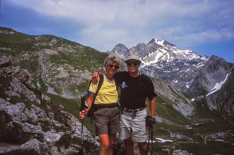 A man and woman with hiking gear stand in front of a hilly background.
