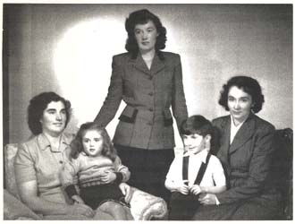 Family portrait with woman standing in middle of two seated women, each holding a small child.