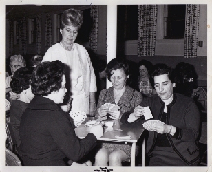 Four women sitting, playing cards, a woman in white standing above. 