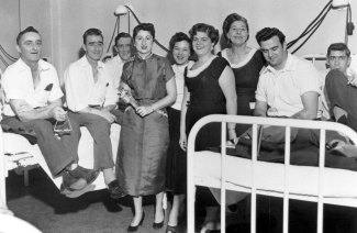 Men sitting on hospital beds and women standing by their side. 