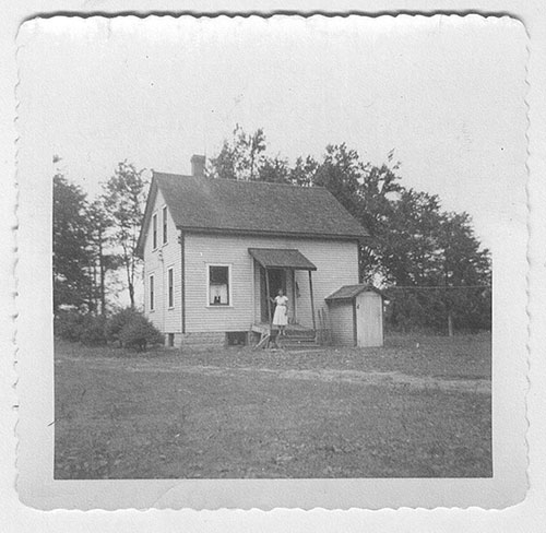 A small farmhouse in the middle of a field, a young girl in a white dress stands on the porch.