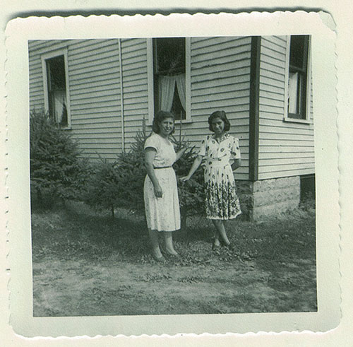Two women stand outside a house, in front of some shrubbery.
