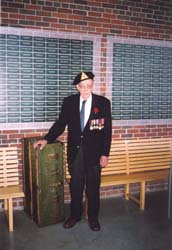 Older Harold, with military medals, standing in front of the Wall of Service at Pier 21.