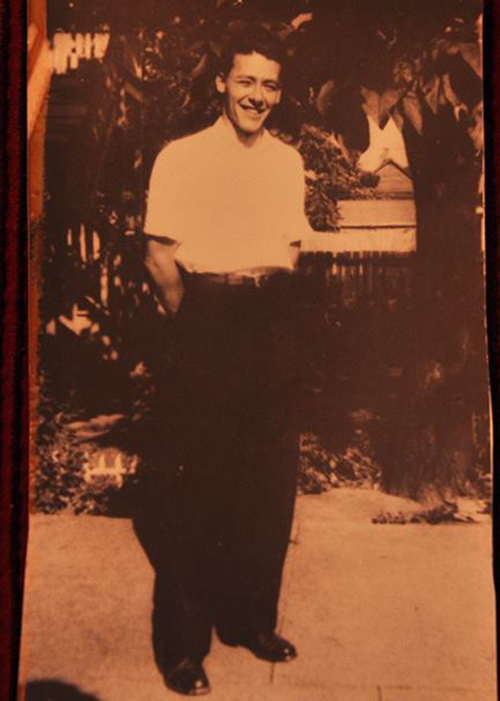 A man stands close to tree, he is wearing a white shirt and has his hands in his pants pockets.