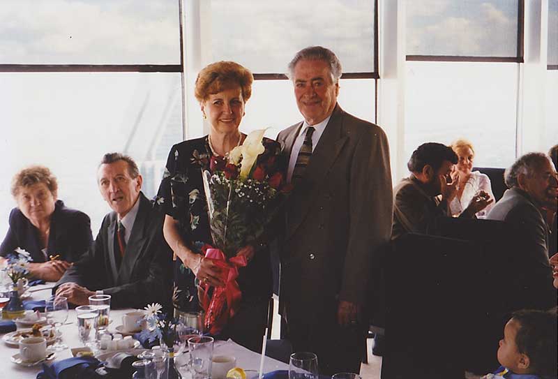 Man and woman standing in front of a table, the woman holds a bouquet of flowers.