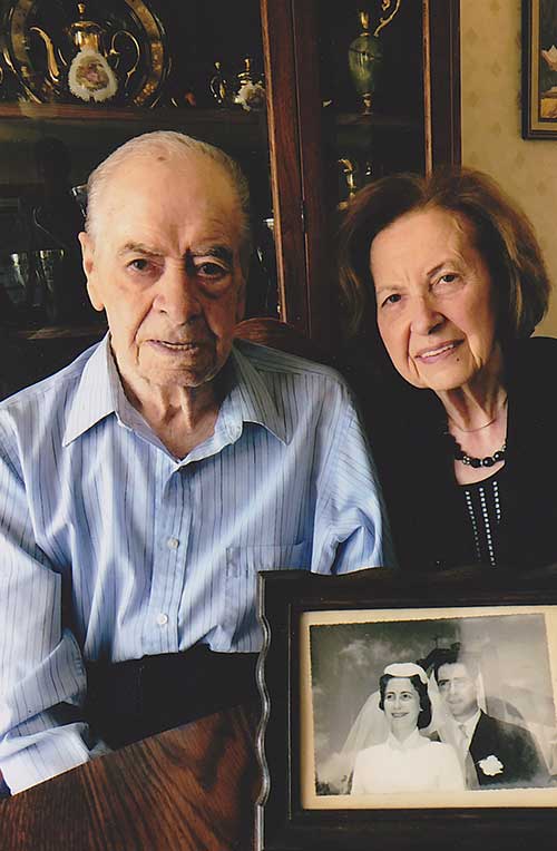 An elderly couple sit behind a framed image of them as a young couple.