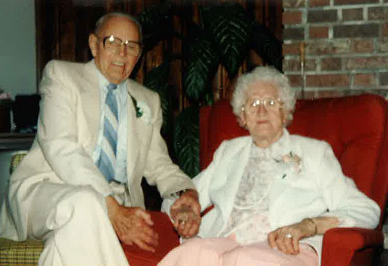 White haired old woman sitting on a red couch and man is sitting next to her and holding her hand.
