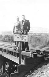 Two young men in suits, standing on railway tracks next to sign reading No trespassing by order.