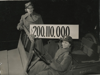 Two female soldiers in an army jeep, holding a sign indicating $200,110,000.