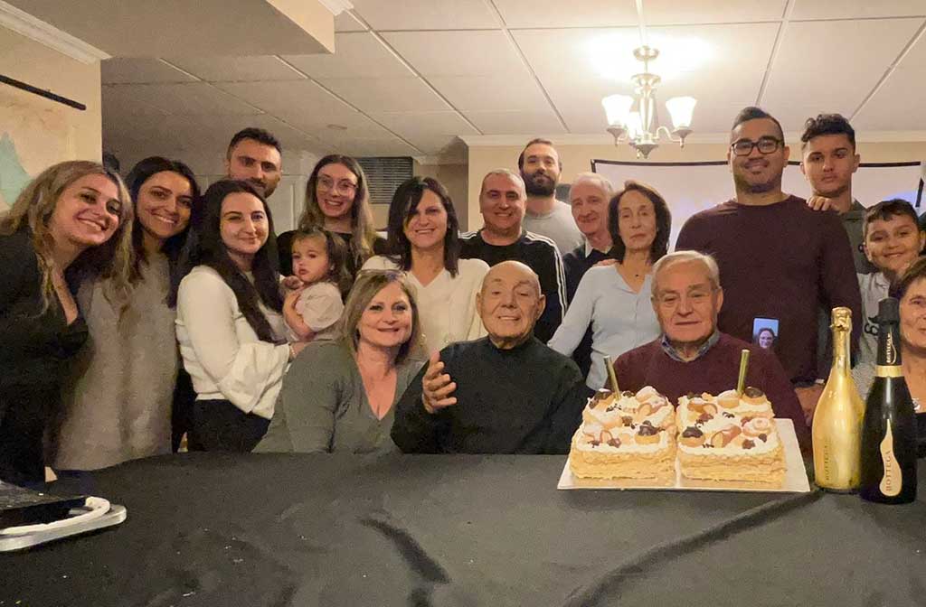 A large family sit around an elderly man, who has a birthday cake in front of him.
