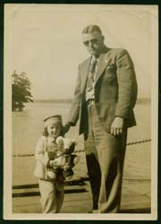 Gerald wearing sunglasses, standing with his young daughter who is holding a teddy bear. 