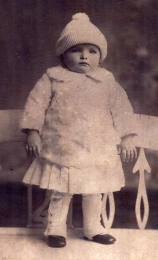 Baby standing, wearing white clothes and a white knitted hat. 