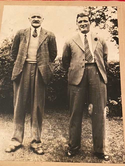 Two men are standing in a garden with trees behind them.