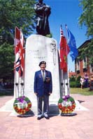 Henry as an older man, standing in front of monument with wreaths and flags next to it.